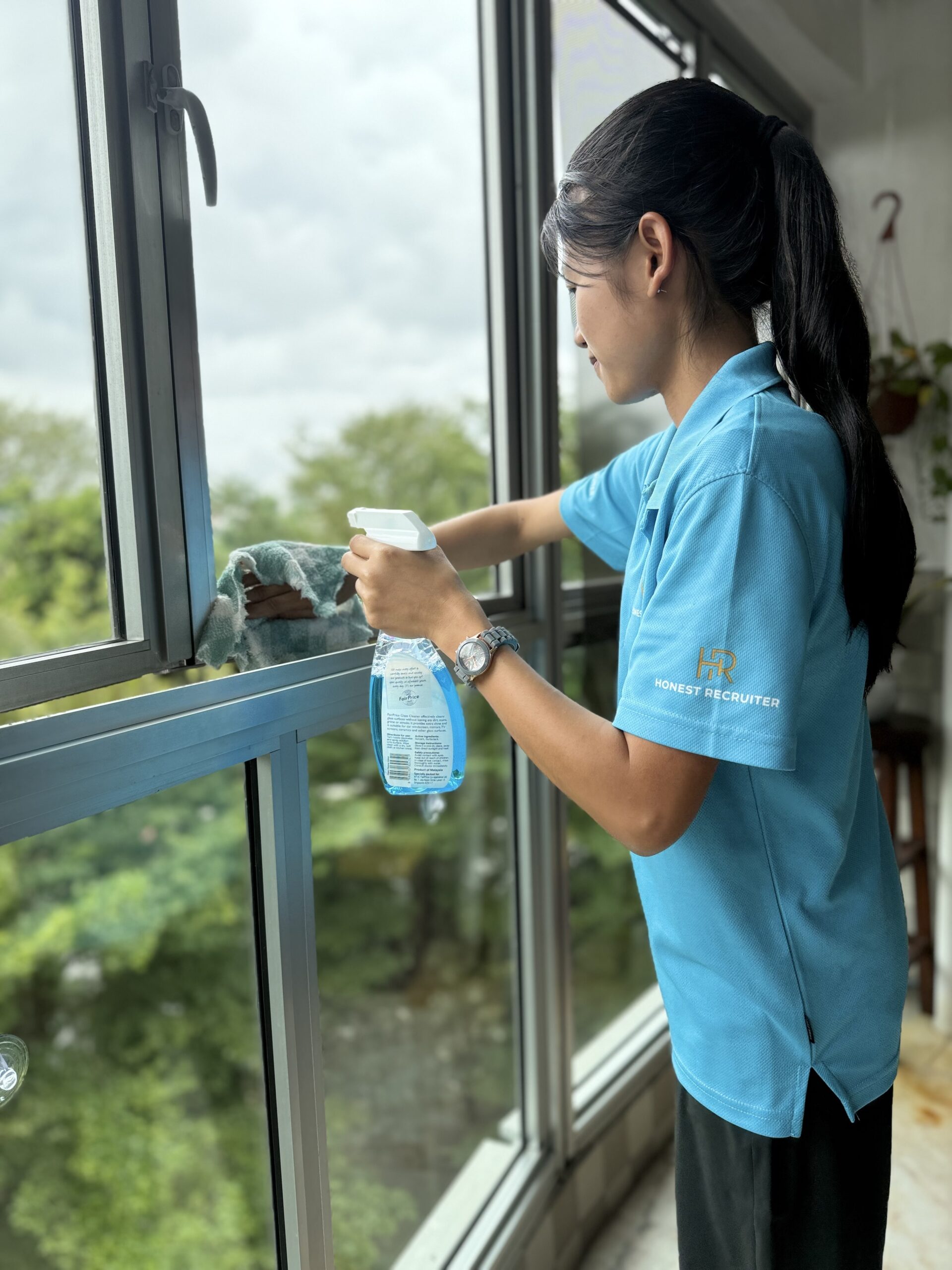Honest Recruiter cleaner wiping a glass window with cleaning spray during home support service in Singapore.